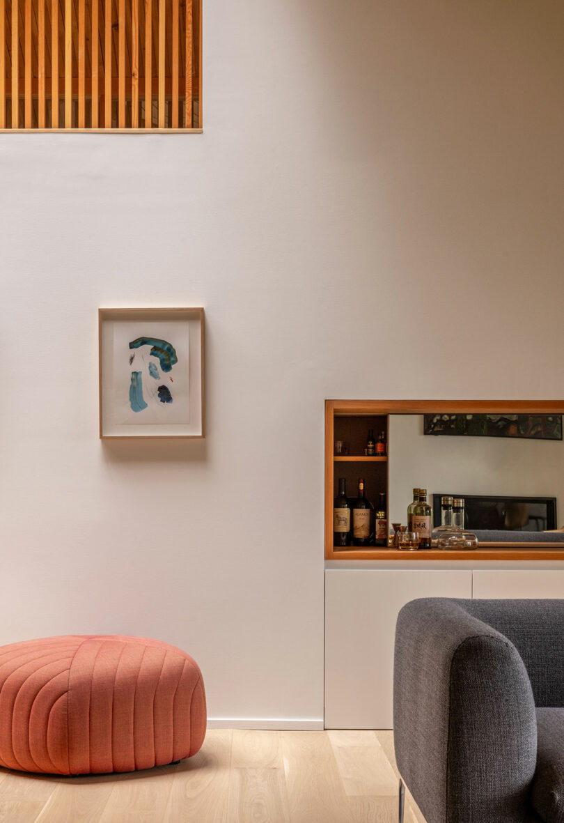 A modern living room with a gray sofa, a pink round ottoman, a minimalist painting on a white wall, and a small bar with bottles visible through a wooden frame cutout.