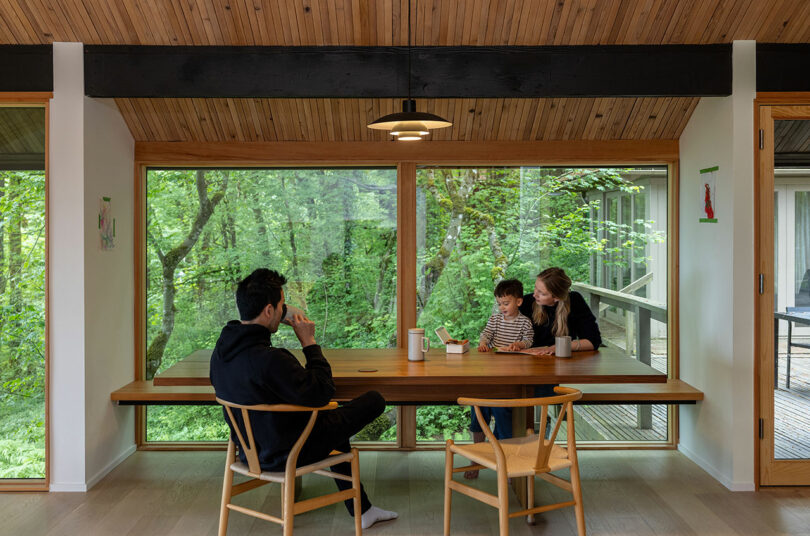 A man, woman, and child sit at a wooden dining table inside a house with large windows showing a forest view.