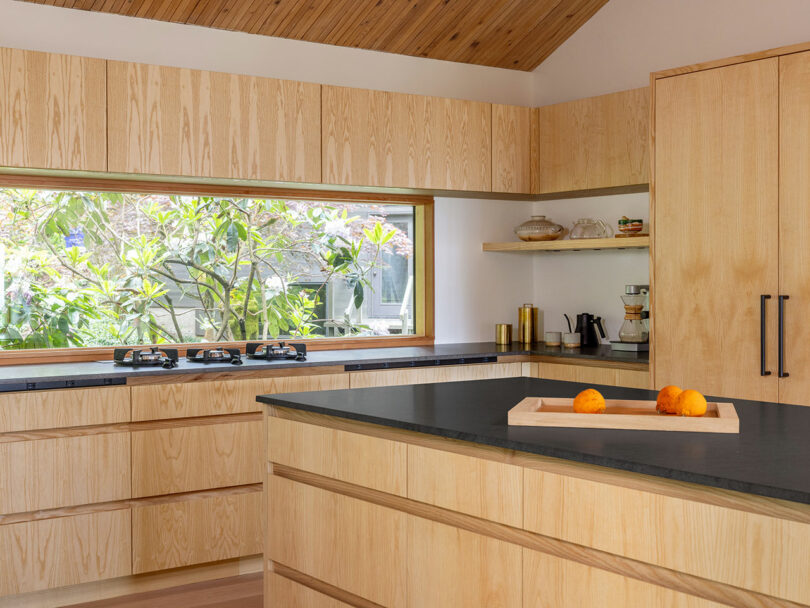 A modern kitchen with light wood cabinets, a black countertop, and a large window. A tray with oranges sits on the island.