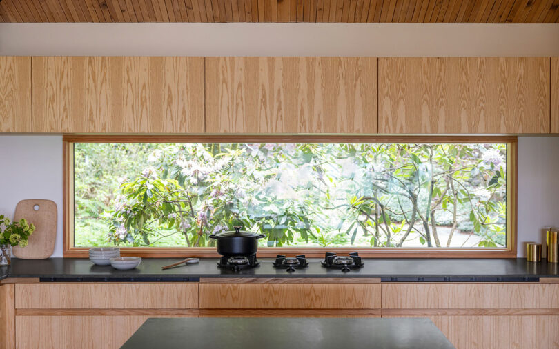 Modern kitchen with wooden cabinets and a large window showcasing green foliage. Black countertop with a stove, pot, and bowls. Wooden ceiling adds warmth to the space.