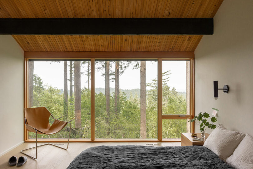 A bedroom with a wooden ceiling, large windows overlooking trees, a bed with dark bedding, a leather chair, a nightstand with a plant, and a pair of slippers on the floor.