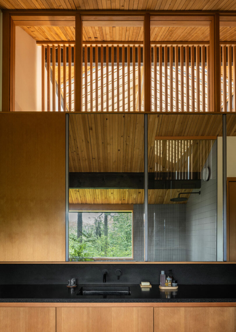 Modern bathroom with wooden cabinets, a black countertop, a large mirror reflecting a wooden ceiling and slats, and a window with a view of green trees.