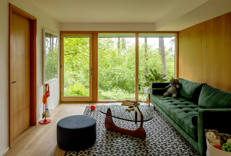 A living room with large glass doors overlooking greenery. It has a green couch, a patterned rug, a round ottoman, a wooden rocking toy, and books on the floor.