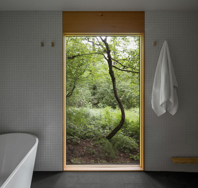 Bathroom interior with a freestanding bathtub, gray tile walls, a towel hanging, and a large window revealing a view of a lush, green forest.