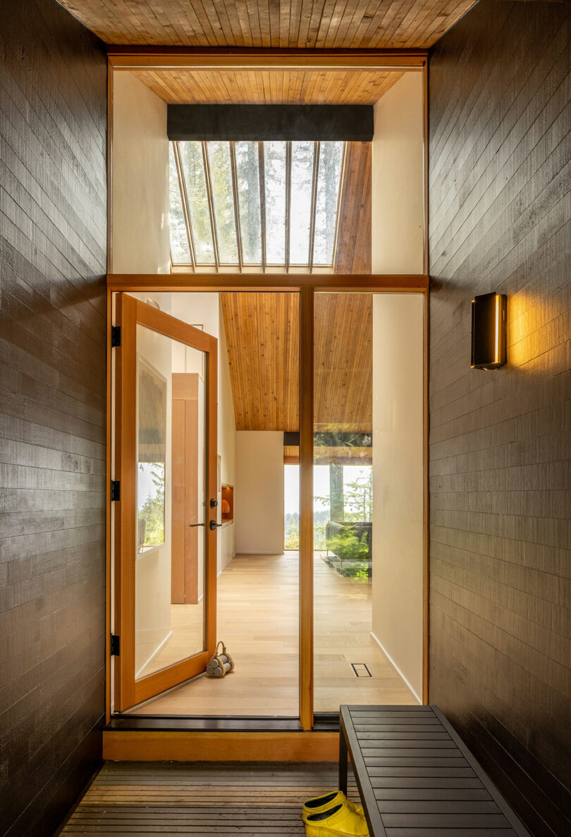 View through a wooden door into a bright interior with wooden ceilings, large windows, and a yellow umbrella on a bench in the entryway.