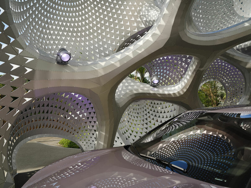 A car is parked under an intricate, geometric metal structure with perforated patterns and embedded lights.