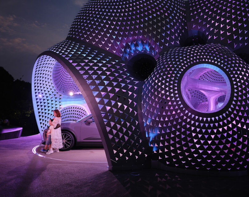 A futuristic building with a textured dome design is illuminated in purple light. A person and a car are partially visible underneath.