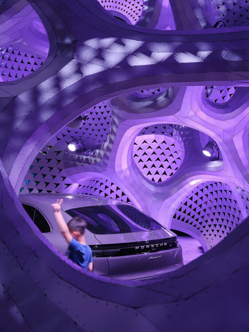 A car with "Porsche" on the back is parked in a futuristic, purple-lit structure. A person stands nearby, partially obscured by the design.