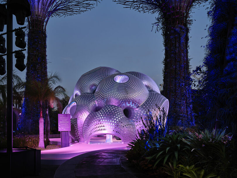 Futuristic dome structure with glowing purple and blue lights surrounded by tall vertical gardens and plants at dusk.