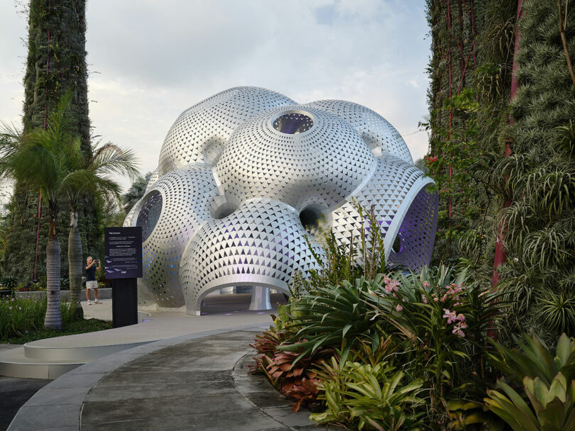 Futuristic dome structure with metallic, bubble-like surface surrounded by lush greenery and palm trees. Pathway leads to entrance.