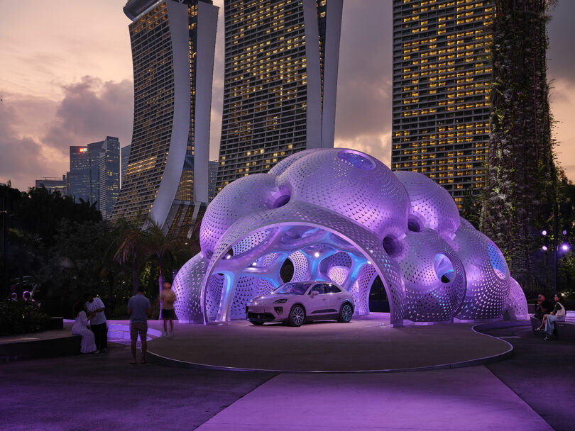 A futuristic car is parked under a purple-lit, dome-like structure. Tall buildings are visible in the background during dusk. People are nearby.