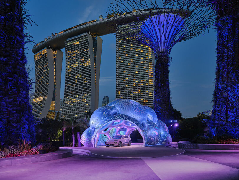 A futuristic car is parked under a glowing, dome-shaped structure with a modern hotel and illuminated trees in the background at dusk.