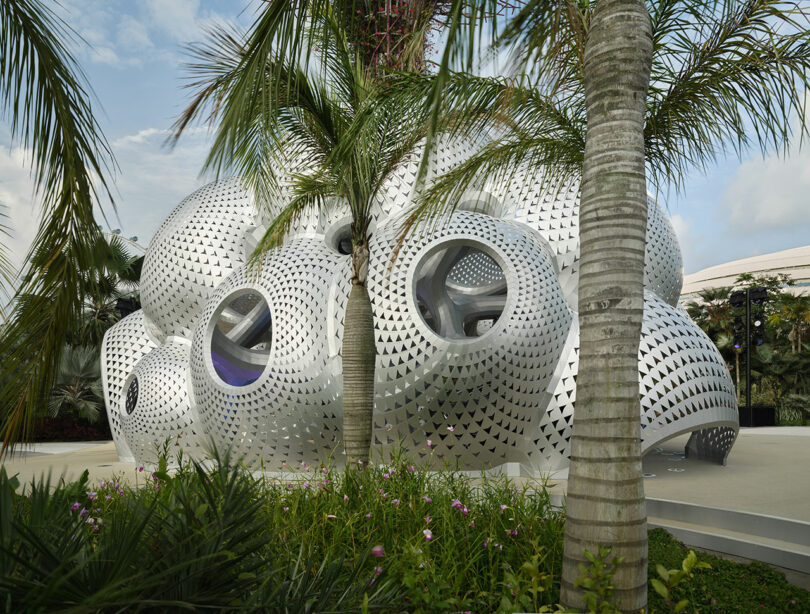 Futuristic silver building with circular windows and perforated panels, surrounded by trees and plants.