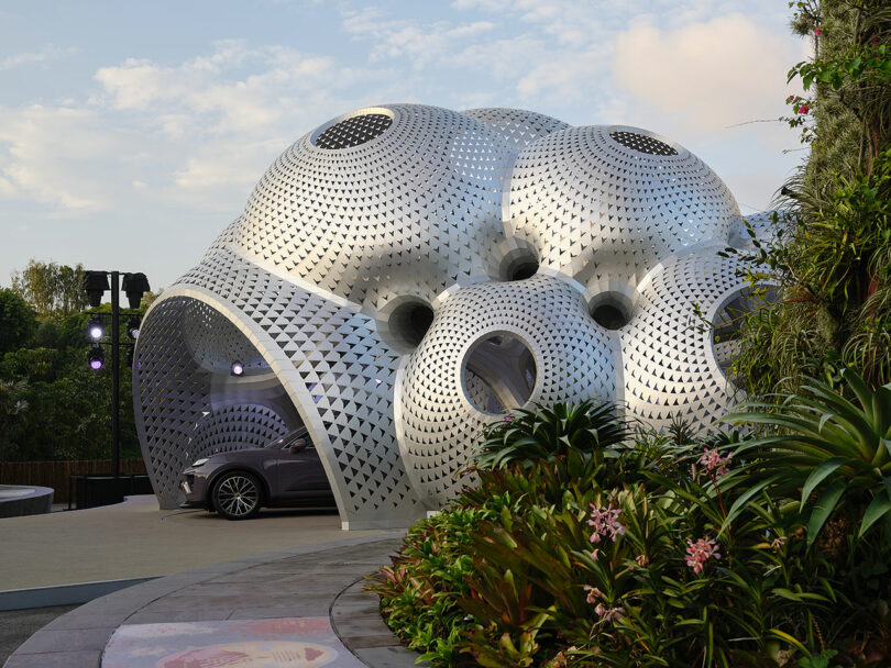 A modern, geometric structure with circular openings stands amidst greenery, partially covering a car.