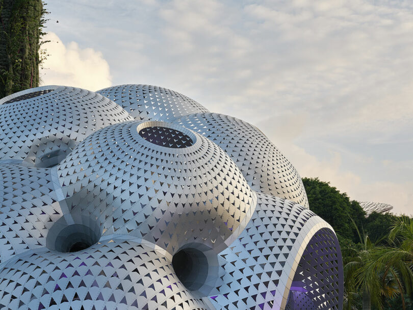 Futuristic silver dome structure with triangular patterns and circular openings, surrounded by greenery.