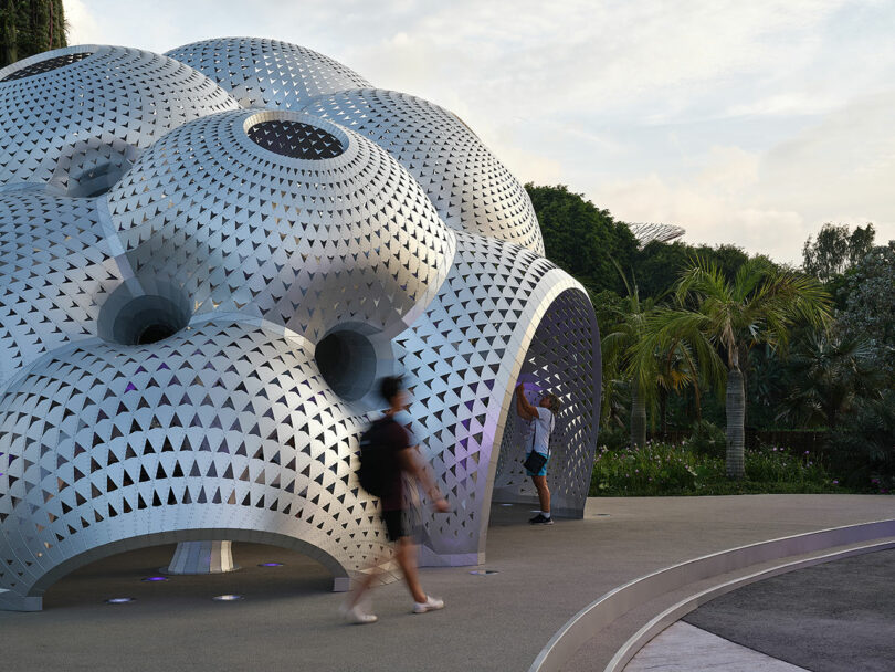 A person walks past and another takes a photo of a futuristic, silver, dome-shaped structure with triangular perforations. Lush greenery surrounds the area.