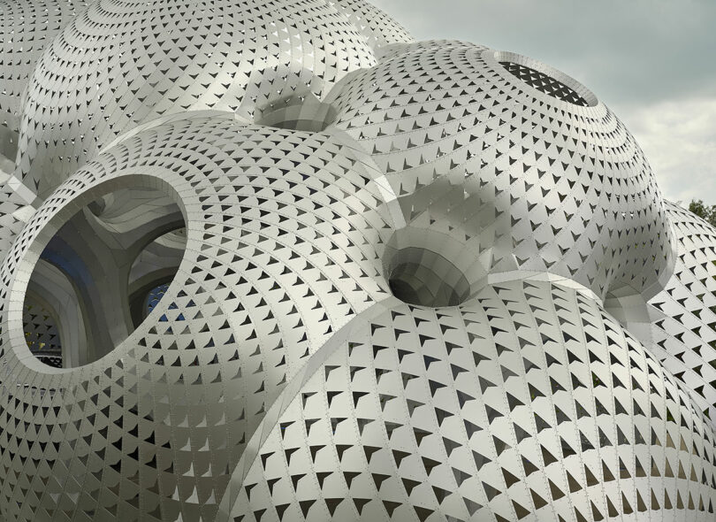 Intricate spherical structure with a pattern of triangular cutouts, featuring multiple large circular openings, under a cloudy sky.