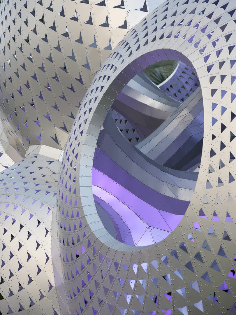 Close-up of a metallic, perforated geometric sculpture with interlocking shapes, featuring a circular opening and illuminated purple accents.