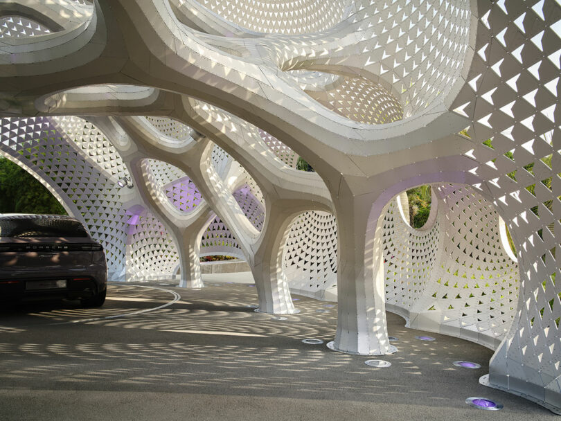 A futuristic, white geometric structure with perforated patterns creates intricate shadows on the ground. A black car is partially visible on the side.