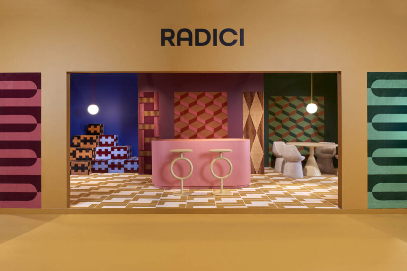 A colorful modern room featuring geometric patterns, a pink bar with two stools, abstract artwork, and a round light fixture. "RADICI" is displayed above the setup.