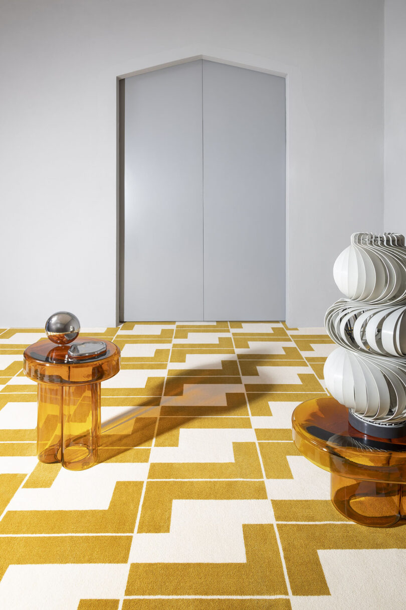 A modern interior with geometric yellow and white flooring, an amber glass side table with a metal sphere, and another table holding abstract white sculptures.