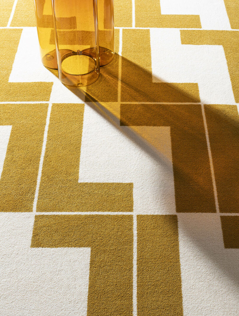 A transparent amber-colored vase casts a shadow on a geometric-patterned rug with mustard yellow and white squares.