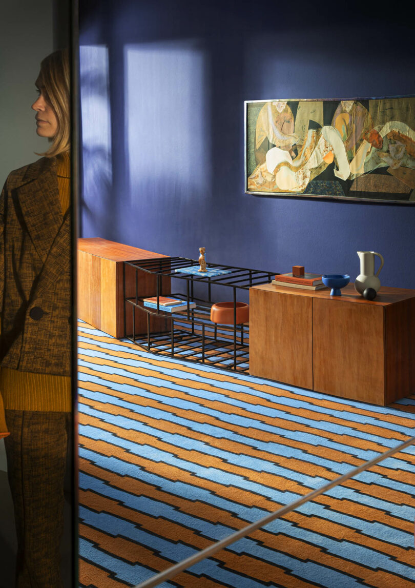 A modern room with blue walls features a geometric carpet, wooden furniture, and abstract art. A person partially appears in the reflection of a mirror on the left.