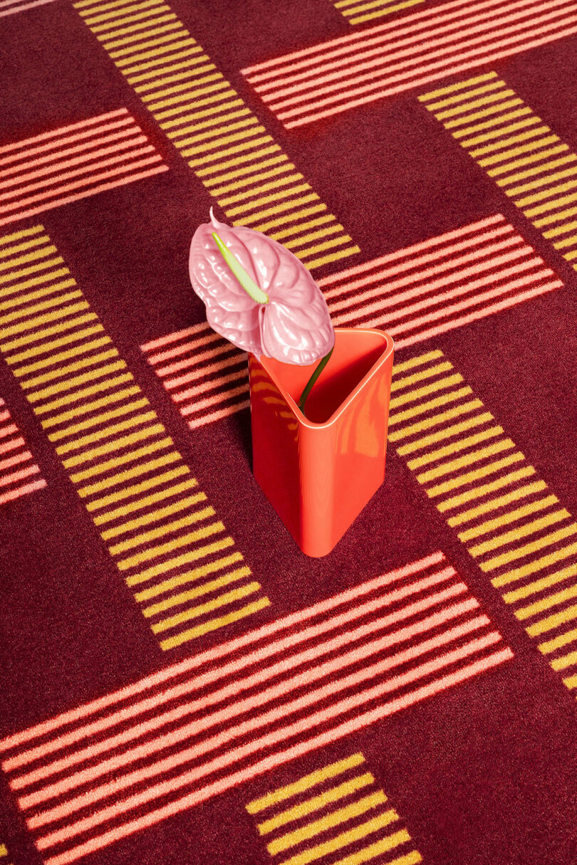 A pink anthurium in a red square vase on a patterned carpet with red, yellow, and beige stripes.