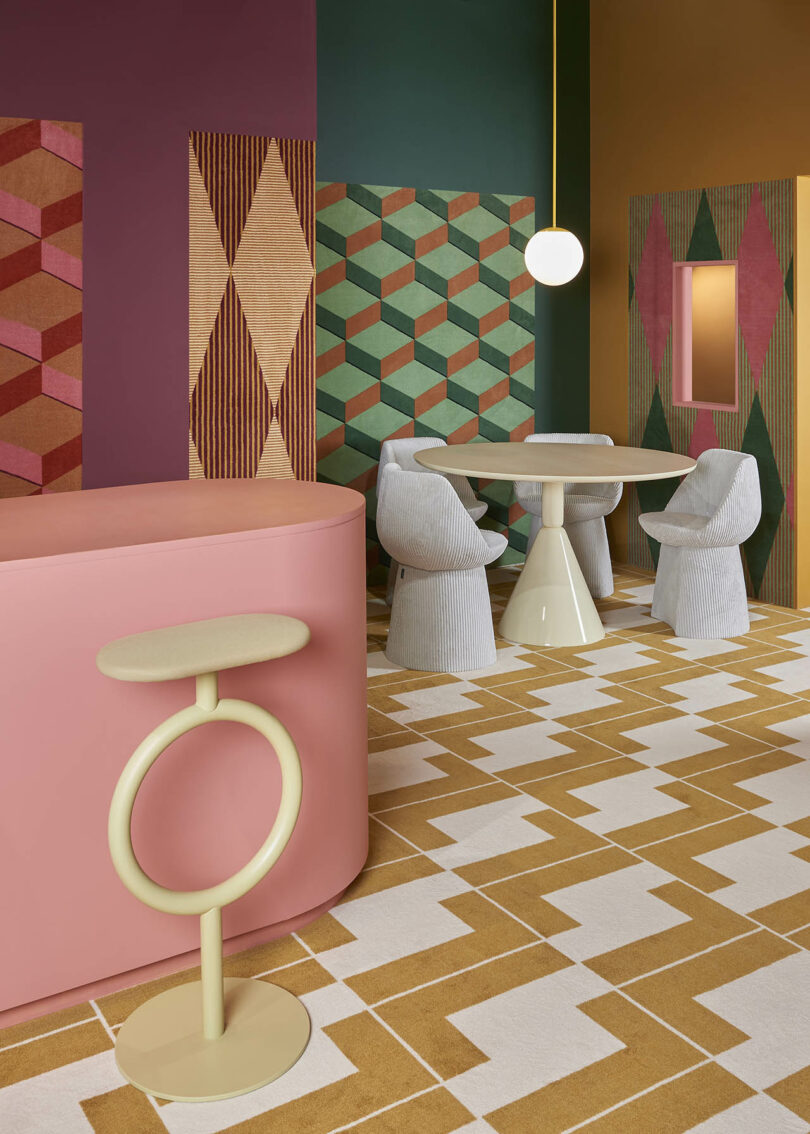 Modern interior with geometric patterns on walls and floor, featuring a round table with four textured chairs and a pink counter with a circular design stool. Overhead, a spherical light hangs.