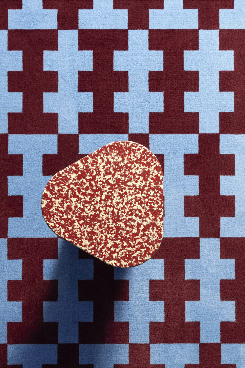 A small, irregularly shaped table with a speckled red and white surface stands on a modern blue and maroon carpet.