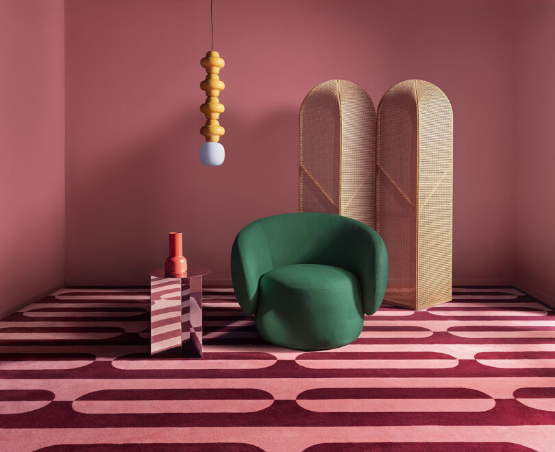 A room with pink walls and geometric carpet features a green chair, a small side table with vases, a modern hanging lamp, and a wicker room divider.