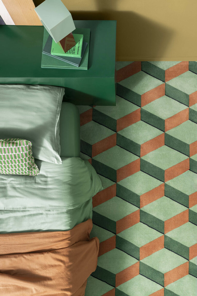 Geometric patterned rug in green and brown, next to a green bed with green and brown bedding, and a green nightstand holding books and a lamp.