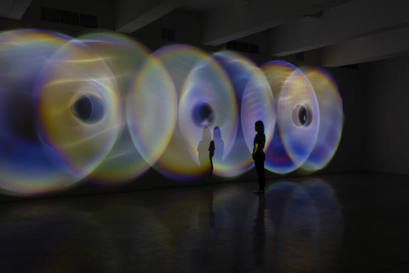 A person stands in a dark room, immersed in a mesmerizing wall display of concentric circles, reminiscent of Olafur Eliasson's ethereal light patterns.