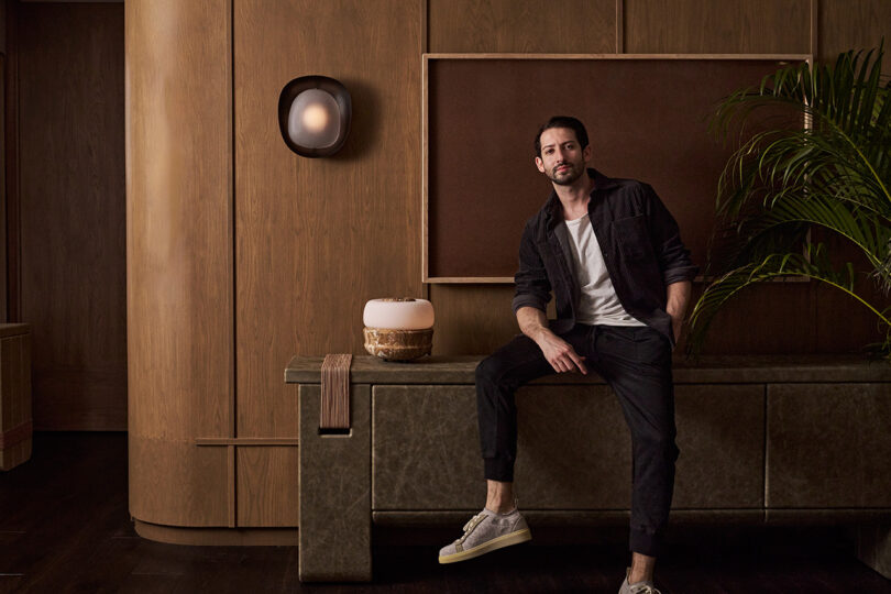 A man sits at a modern console in a stylish room with wooden walls, a potted plant, and wall art, illuminated by a Pell Chandelier that adds an elegant touch to the ambiance.