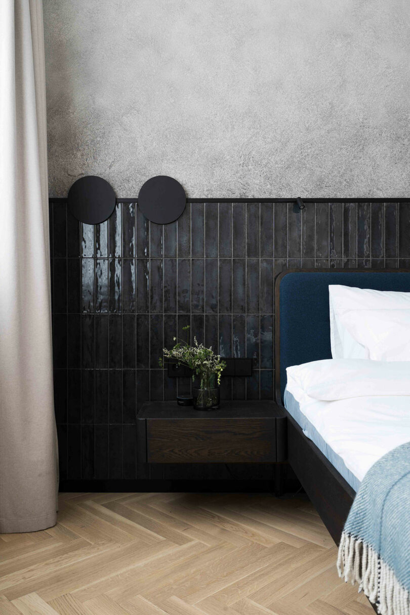 Minimalist bedroom with a dark tiled wall, a black nightstand holding a plant, and a neatly made bed. Light wood herringbone floor and white curtain on the left.