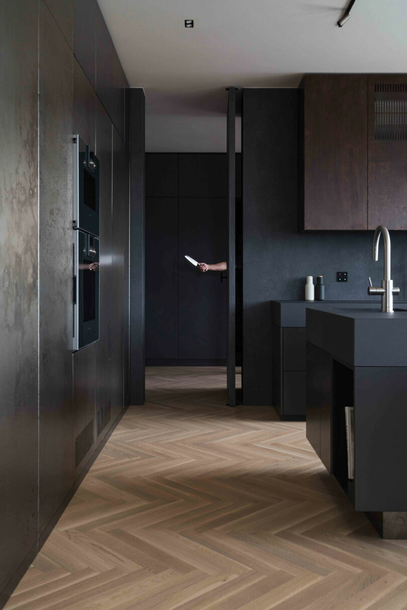 A modern kitchen with dark cabinetry, wood flooring, and a person holding a knife partially visible through an open doorway.