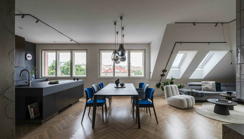 A modern open-plan living space with a dining table, blue chairs, a kitchen area, and a cozy seating area. Large windows and skylights provide natural light.