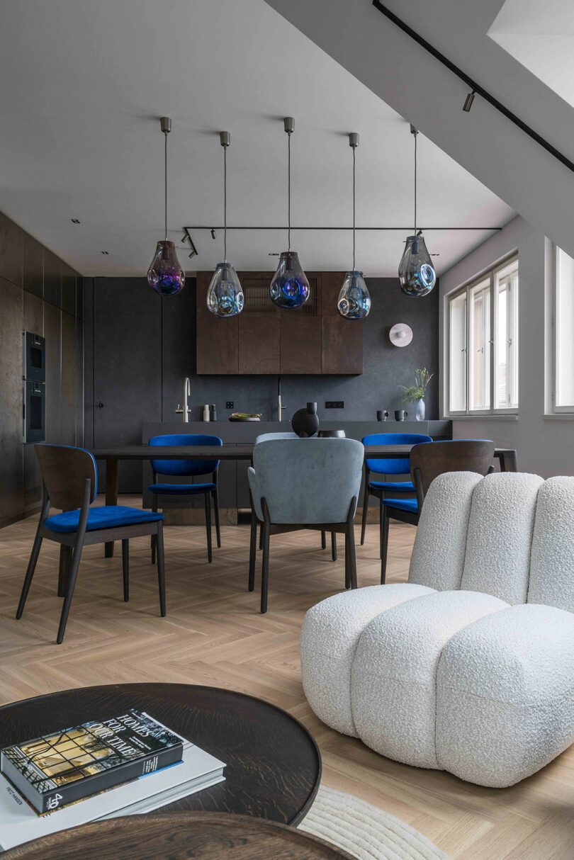 Modern living room with a plush white chair, wooden round table with books, and a dining area featuring blue chairs and pendant lights. Brown cabinets are seen in the background.