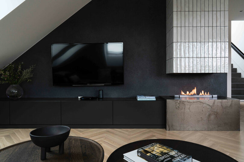 Modern living room with dark wall, wall-mounted TV, minimalist fireplace, black cabinetry, and wood flooring. Stairs in the background; round coffee table with books and black bowl.