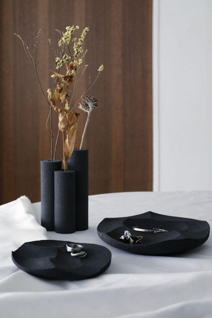 A minimalist arrangement with dried flowers in a black three-part vase and black geometric plates holding jewelry on a white cloth-covered table.