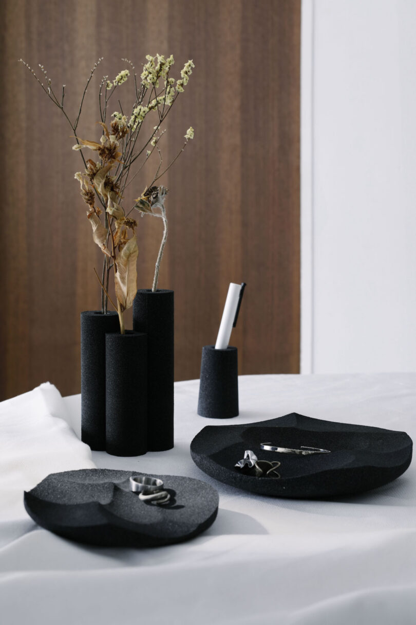 Black ceramic vases with dried flowers and a pen rest on a table. Two black dishes hold silver rings and earrings