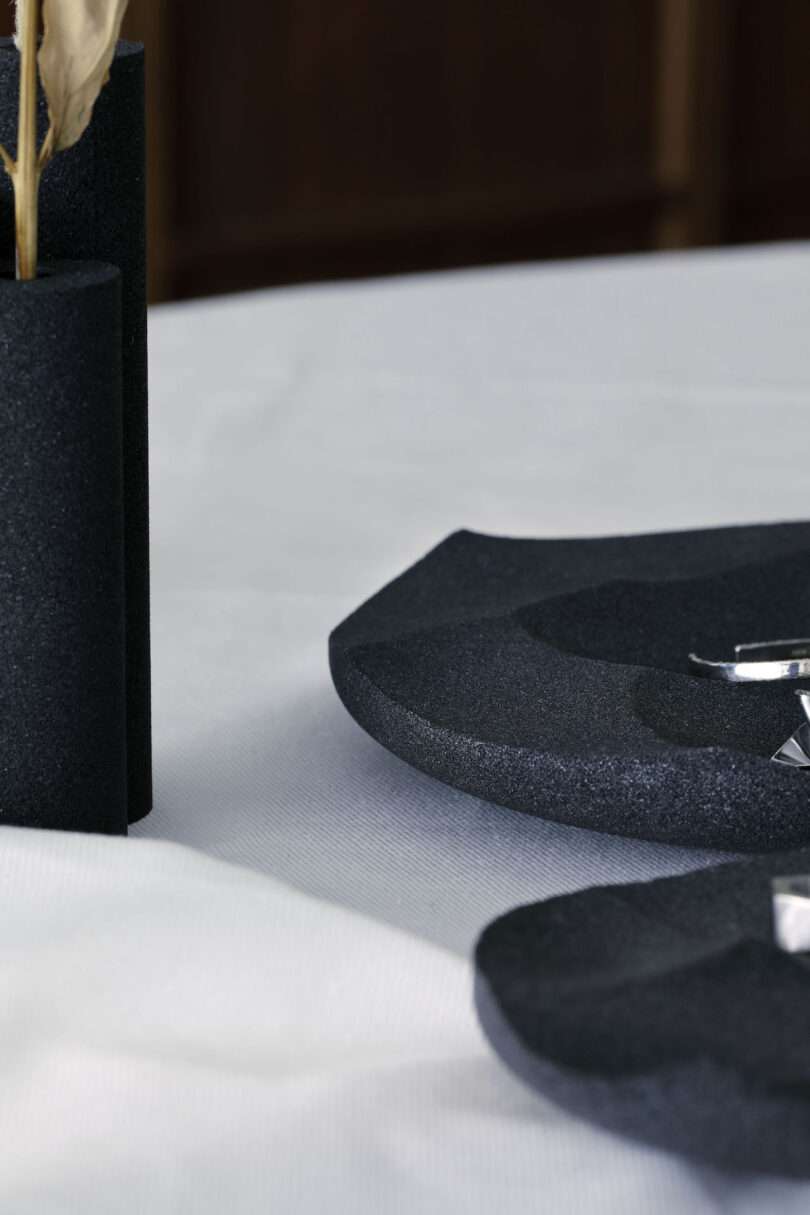 Close-up of black textured ceramic plates and a cylindrical vase on a white tablecloth, holding a dried plant stem