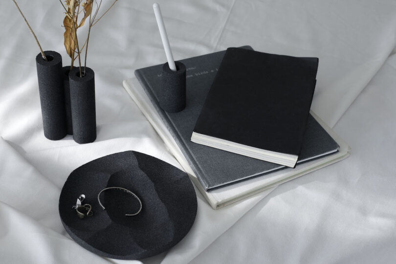 A neatly arranged scene with black stationery, a notebook, a pen holder, two vases with dried plants, and a dish holding a bracelet and earrings on a white fabric surface`