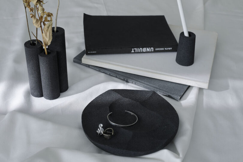 A black bowl with jewelry, a black and white book stack, black cylindrical vases with dried flowers, and a white candle on a white cloth