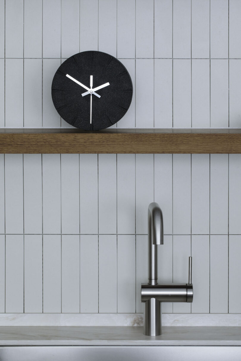 A black wall clock with silver hands sits on a wooden shelf above a stainless steel faucet and sink against a white tiled wall