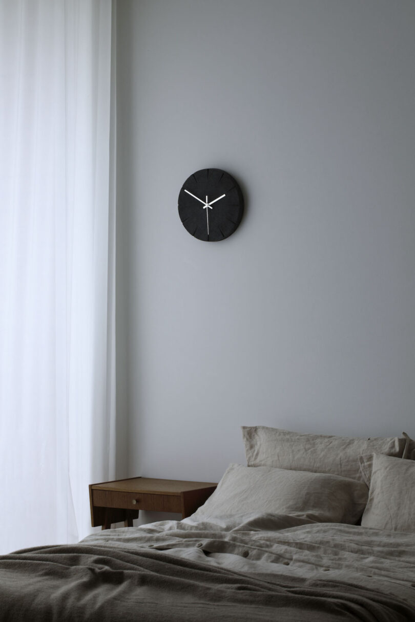 Minimalist bedroom scene with a black wall clock, white curtains, a wooden side table, and a neatly made bed with neutral bedding