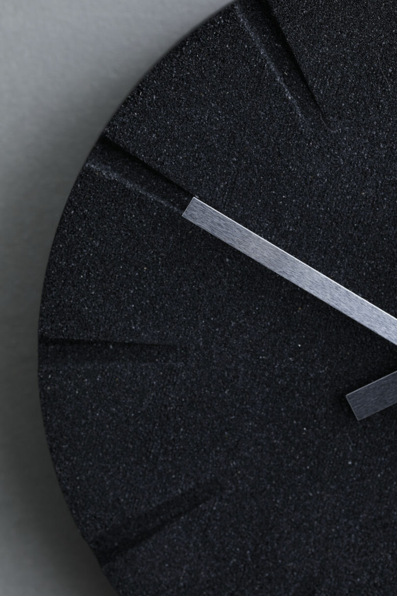 Close-up of a minimalist black wall clock with silver hands. Only a portion of the clock face is visible, featuring subtle hour markers