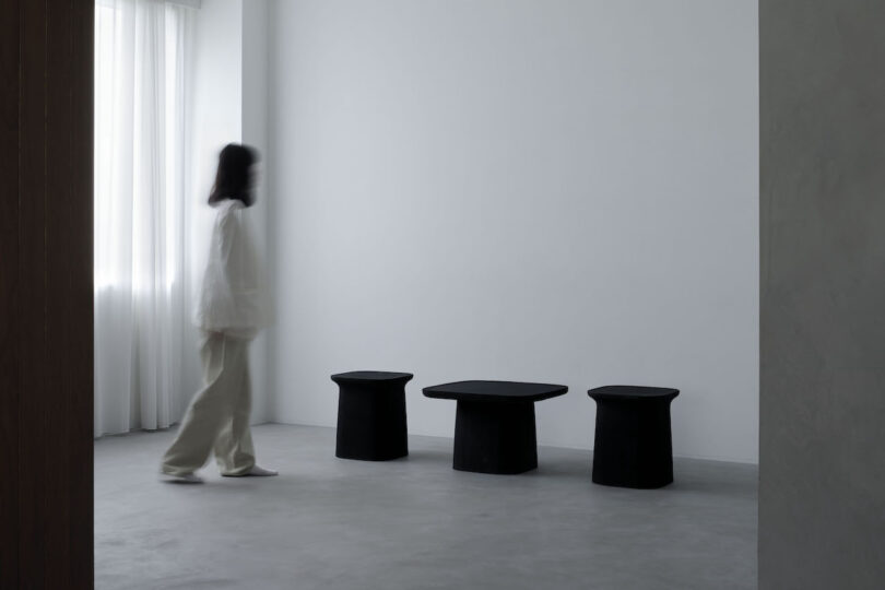 Blurry image of a person in a white outfit walking past three black minimalist stools in a sparsely furnished, light-filled room.