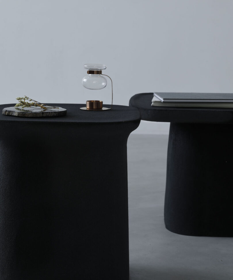 Minimalist interior featuring two black tables. One holds a decorative plant and a small glass hourglass lamp, while the other has stacked books.