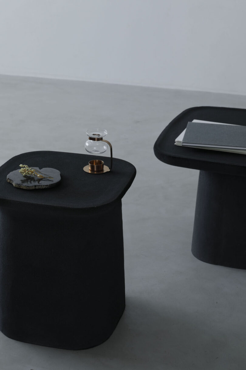 Two black, square tables on a gray floor; one holds a small glass and copper object, and the other has books and a slate with a plant